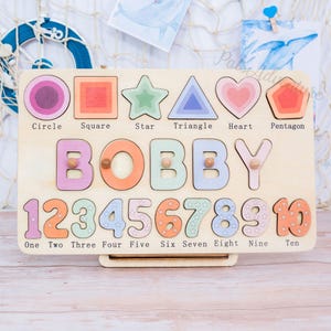 May include: Wooden puzzle board with colorful shapes, the name "BOBBY", and numbers 1-10. Shapes include a circle, square, star, triangle, heart, and pentagon. Each shape and number is a separate piece. The board is light brown.