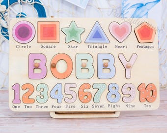 May include: Wooden puzzle board with colorful shapes, the name "BOBBY", and numbers 1-10. Shapes include a circle, square, star, triangle, heart, and pentagon. Each shape and number is a separate piece. The board is light brown.