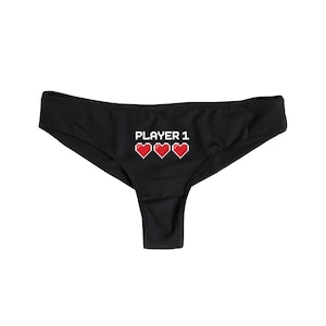 以下が含まれることがあります： 「PLAYER 1」の文字と3つの赤いピクセル化されたハートがプリントされた黒い下着。ローライズカットで、柔らかい素材で作られています。