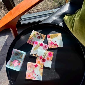 Puede incluir: Un conjunto de posavasos cuadrados de resina con flores secas y detalles de pan de oro. Los posavasos presentan arreglos florales rosas, rojos y blancos sobre un fondo azul claro. Se exhiben sobre una bandeja redonda negra.