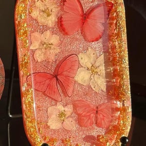 以下が含まれることがあります： ピンク色のベースに赤い蝶と白い花が埋め込まれた、楕円形の樹脂トレイ。トレイの縁は金色のフレークで飾られており、装飾的で芸術的な作品となっています。