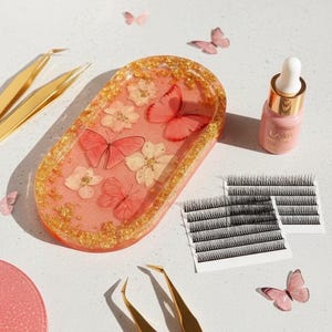 Op de afbeelding: Esthetische opstelling met een roze harsbakje met vlinders en gedroogde bloemen, een gouden pincet, wimperserum en valse wimpers. Het bakje heeft een gouden glitterrand. Zachte verlichting en vlinderaccenten.