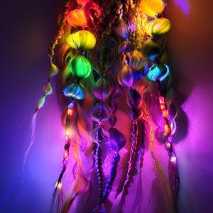 Puede incluir: Un vibrante accesorio para el cabello multicolor con orbes brillantes y mechones trenzados. La pieza muestra un espectro de colores, incluyendo amarillo, verde, azul y morado, creando un efecto luminoso. El diseño es probablemente para un look festivo o de actuación.