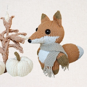May include: A knitted fox with a scarf, next to knitted pumpkins and a corn stalk. The fox is brown, white, and tan, with a black nose and large eyes. The pumpkins are white, and the corn stalk is tan.