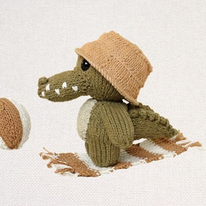May include: A green knitted crocodile wearing a brown knitted hat and holding a brown and white knitted ball. The crocodile is sitting on a brown and white striped knitted rug.