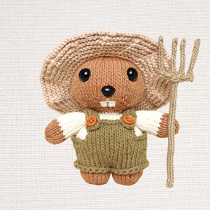 May include: A knitted toy beaver wearing a brown hat and overalls, holding a brown stick with a fork on the end.