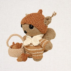 以下が含まれることがあります： 茶色のニットのリスのぬいぐるみが、茶色のどんぐり帽子とストライプのセーターを着ています。リスは、中に2匹の小さな茶色のニットのリスが入ったバスケットを持っています。