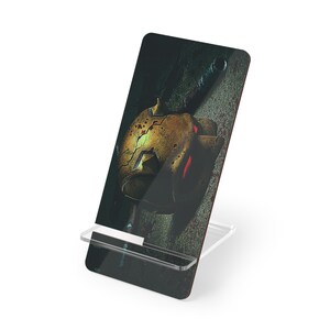 Op de afbeelding: Een telefoonstandaard met een digitale print van een beschadigde, goudgele helm met rode accenten en een donkere achtergrond. De standaard is gemaakt van helder acryl.