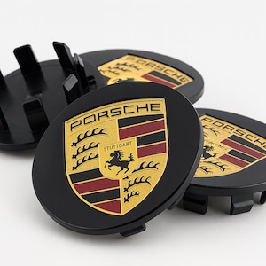 May include: Four black wheel centre caps featuring the Porsche logo. The logo includes a gold shield with a black horse, red and black stripes, and the word "PORSCHE" in gold. The caps are made of plastic.