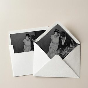 May include: Two white envelopes, one open, displaying black and white photographs of a couple. The open envelope reveals a full image of the couple, while the other shows a partial view. The background is a neutral beige.