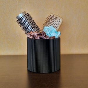 May include: A black, vertically-lined cylindrical container holding a round brush, a wooden paddle brush, and silk scrunchies in brown and blue. The container sits on a dark brown surface against a light brown background.