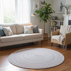May include: A round, braided rug in a living room setting. The rug is primarily light gray with subtle pink accents, creating a soft, textured appearance. The rug is placed on a hardwood floor, complementing the neutral tones of the room's furniture and decor.