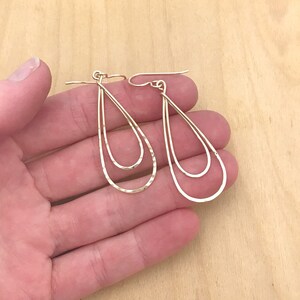Peut inclure: Une paire de boucles d'oreilles dor&eacute;es en forme de goutte d'eau. Chaque boucle d'oreille pr&eacute;sente un design &agrave; double couche, avec une texture martel&eacute;e sur la couche ext&eacute;rieure. Les boucles d'oreilles sont pr&eacute;sent&eacute;es sur une surface en bois clair.