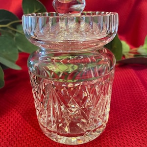 May include: A clear, cut-glass jar with a lid and a small, round knob. The jar has a diamond pattern and sits on a red surface. The lid is a separate piece with a ribbed edge.