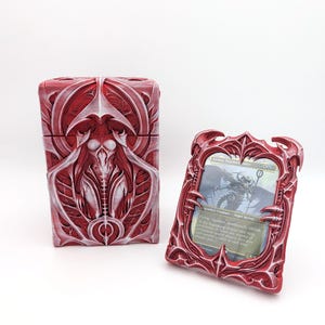 May include: A red and white rectangular deck box with intricate biomechanical relief patterns and a matching red frame holding a trading card titled Athreos, God of Passage, featuring a dark creature illustration and printed game text.
