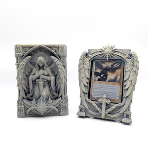 May include: Two grey, intricately detailed card holders. One features an angel with a sword, the other holds a card with an angel illustration. Both have sun and wing motifs, suggesting a fantasy or collectible theme.