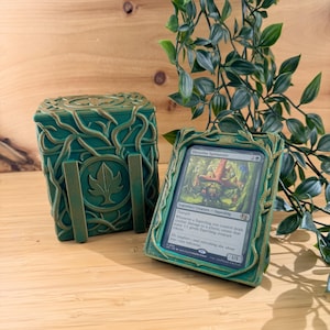 May include: Green card holder and deck box with a leaf design. The card holder displays a card with the text "Shmoofus Sproutfire". The deck box and card holder are made of a textured material and sit on a wooden surface.