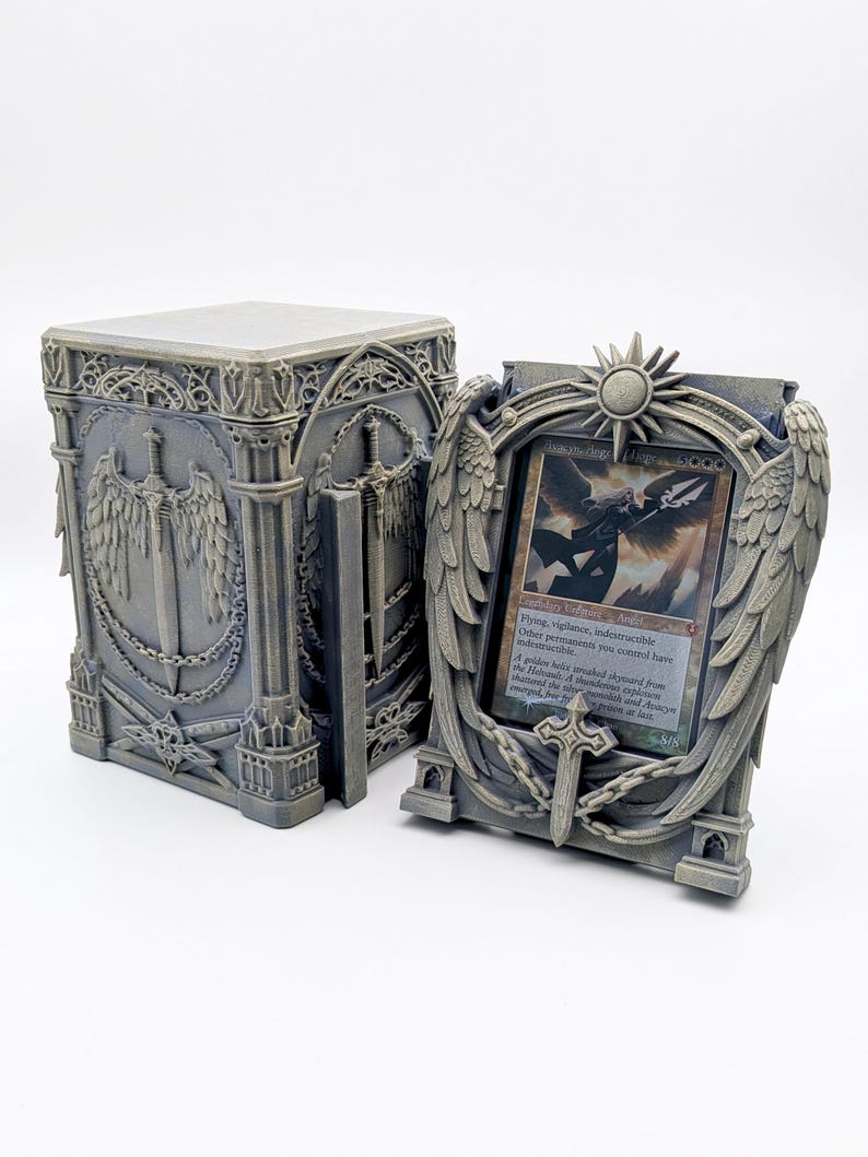 May include: A gray, gothic-style card holder and box set. The box features engraved swords, wings, and chains. The card holder has a sun and wing design, holding a card with text. The set is made of a durable material.