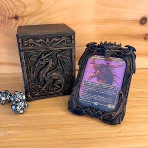 May include: A dark brown dragon-themed card holder and matching box. The card holder displays a Magic: The Gathering card, "The Ur-Dragon." Dice are visible in the foreground. The box and holder feature dragon relief carvings.