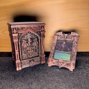 Pode incluir: Uma caixa e moldura para cartas em estilo gótico, cor de cobre. A caixa apresenta uma criatura semelhante a um morcego com uma cruz num escudo. A moldura tem um design de caveira e asas de morcego.