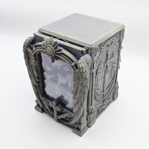 May include: A gray, rectangular card storage box with ornate, gothic-style detailing. The box features a hinged lid and a front panel with a framed display area. Decorative elements include wings, sunbursts, and swords.