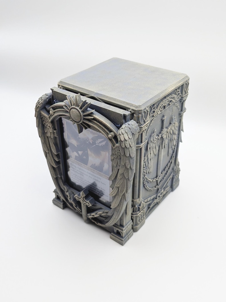 May include: A gray, rectangular card storage box with ornate, gothic-style detailing. The box features a hinged lid and a front panel with a framed display area. Decorative elements include wings, sunbursts, and swords.