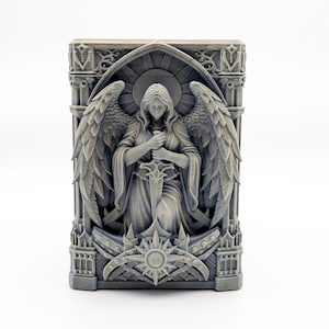 May include: A gray, rectangular sculpture of an angel holding a sword, with large detailed wings. The angel is set within a gothic-style frame with ornate detailing and a sunburst design at the base. The sculpture is set against a white background.