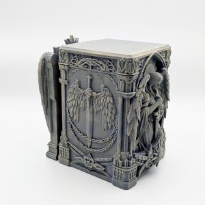 May include: A gray, gothic-style box with detailed relief carvings. The box features angel figures, a sword with wings, and ornate architectural elements. The design is intricate and the overall aesthetic is reminiscent of a medieval cathedral.