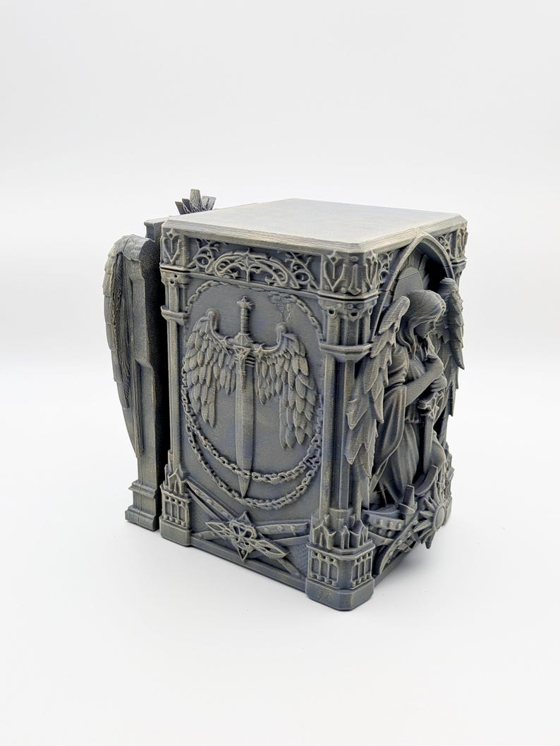 May include: A gray, gothic-style box with detailed relief carvings. The box features angel figures, a sword with wings, and ornate architectural elements. The design is intricate and the overall aesthetic is reminiscent of a medieval cathedral.