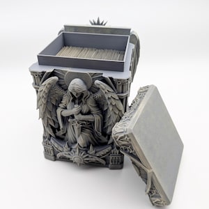May include: A gray, rectangular box with an open lid, featuring an angel sculpture on the front. The box has a detailed design with wings and a halo. The box is designed to hold small items.