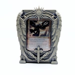 May include: A gray card holder with angel wings and a sun design, holding a Magic: The Gathering card. The card features an angel illustration and the text "Avacyn, Angel of Hope".