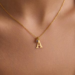 May include: A gold-colored necklace with a delicate chain. The pendant features the letter "A" in a serif font, with a small, round, purple gemstone above it. The necklace is worn against a light blue top.