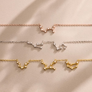 Peut inclure: Trois colliers délicats avec des motifs de constellations. Les colliers sont en or rose, argent et or. Chaque collier a une chaîne et un pendentif constellation orné de petites pierres scintillantes. Le fond est de couleur neutre.