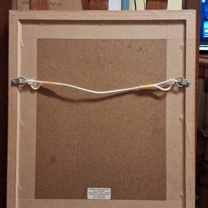 May include: Back of a framed picture, showing the mounting hardware. The frame is light brown, with a brown backing. A white cord is strung between two metal eyelets for hanging. The frame is resting on a wooden chair.