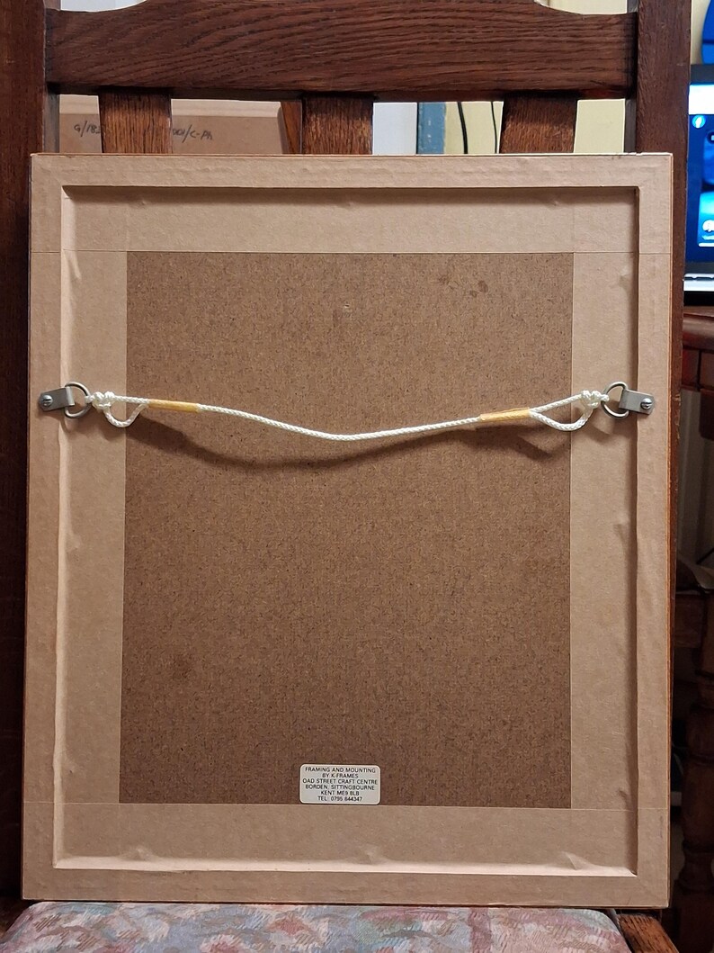 May include: Back of a framed picture, showing the mounting hardware. The frame is light brown, with a brown backing. A white cord is strung between two metal eyelets for hanging. The frame is resting on a wooden chair.