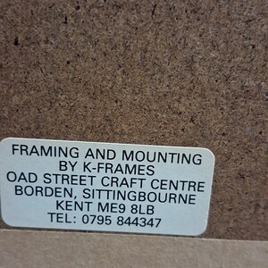 May include: A close-up of a brown textured surface with a white rectangular label. The label reads "FRAMING AND MOUNTING BY K-FRAMES OAD STREET CRAFT CENTRE BORDEN, SITTINGBOURNE KENT ME9 8LB TEL: 0795 844347".