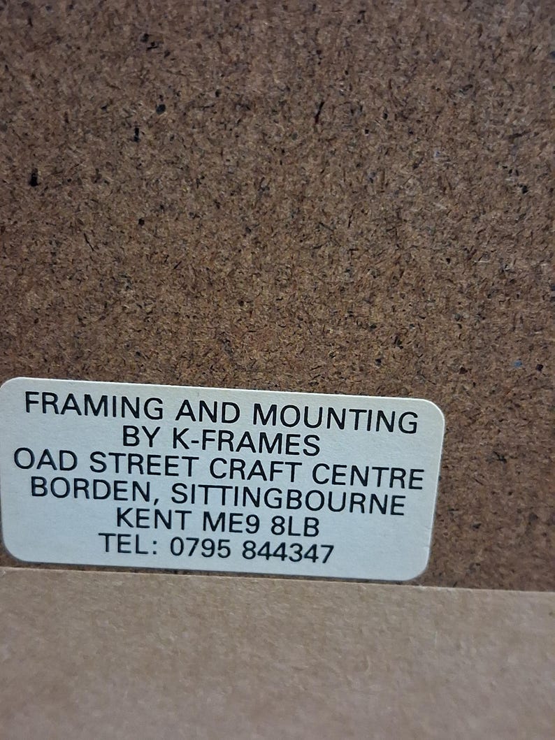 May include: A close-up of a brown textured surface with a white rectangular label. The label reads "FRAMING AND MOUNTING BY K-FRAMES OAD STREET CRAFT CENTRE BORDEN, SITTINGBOURNE KENT ME9 8LB TEL: 0795 844347".