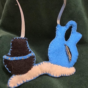 May include: Handmade felt ornament in blue, black, and cream colours. The ornament features a stylized design with a feather charm. The edges are stitched with contrasting thread, and a silver ribbon is attached for hanging. The ornament is approximately 10 cm long.