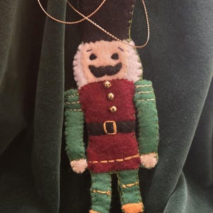Pode incluir: Enfeite de Natal de feltro feito à mão em forma de quebra-nozes. O enfeite apresenta um chapéu preto, um colete vermelho com botões dourados, mangas e calças verdes e botas pretas. Uma corda dourada é anexada para pendurar.