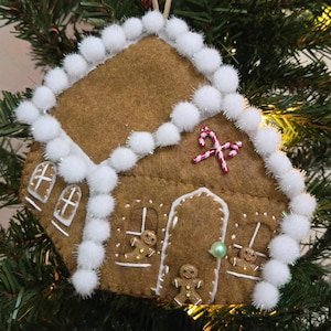 Könnte beinhalten: Ein brauner Lebkuchenhaus-Weihnachtsschmuck aus Filz mit weißem Bommelbesatz. Der Schmuck hat weiße Zuckergussdetails, Lebkuchenmännchen und Zuckerstangen-Akzente. Er hängt an einem Weihnachtsbaum.