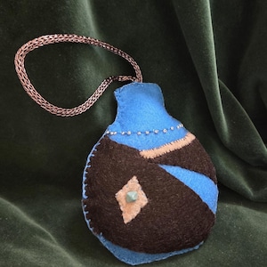 May include: A handmade felt pouch in shades of blue and brown, with a decorative geometric design and a small, square, turquoise-colored gem. The pouch is attached to a chain. The background is a dark green velvet.