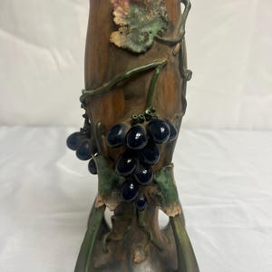 Puede incluir: Un jarrón marrón con superficie texturizada, con un diseño de uvas y hojas. Las uvas son de color azul oscuro y las hojas verdes y rosas. El jarrón tiene una base acampanada y forma cilíndrica.