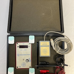 Op de afbeelding: Een open zwarte koffer met een röntgenpulsteller, röntgensensor, kabels en sondes. De pulsteller is wit en rood met een digitaal display en de tekst "Engineered Systems & Designs". De röntgensensor is geel met een doelwit.