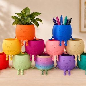 May include: A pyramid of eleven colourful, round, smiling planters with dangling legs. Each pot features embossed text including phrases like 'Thank You', 'Best Teacher', and 'Super Teacher'. One orange pot holds a green plant, and a blue pot holds pens.