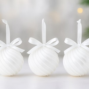 May include: Three spherical white baubles with a swirling ribbed texture, each topped with a matching white satin ribbon tied into a bow and a loop for hanging. The baubles have a smooth, matte finish and are arranged in a horizontal row.