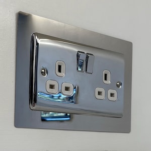 May include: A shiny, chrome-finished electrical outlet with two sockets and two switches. The outlet is mounted on a rectangular plate and installed on a white wall. The design is modern and sleek, reflecting light.