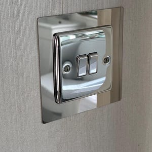 Puede incluir: Un interruptor de luz doble cromado pulido sobre una placa cuadrada. La placa está montada en una pared gris claro texturizada. El interruptor tiene dos interruptores basculantes rectangulares. La pared tiene un patrón de rayas verticales.