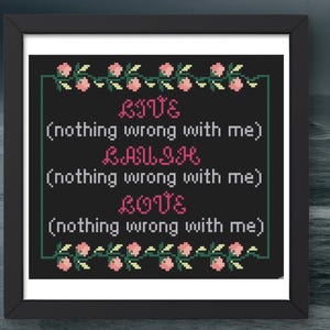 May include: A framed cross-stitch artwork with the words "LIVE, LAUGH, LOVE" in pink, with the phrase "(nothing wrong with me)" below each word. The artwork is framed in a black frame and decorated with pink flowers and green leaves.