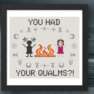May include: A framed cross-stitch embroidery on light fabric featuring a black horned figure and a person in a pink dress standing by an orange fire. The text reads YOU HAD YOUR QUALMS?! with small grey symbols arranged in a circle around them.