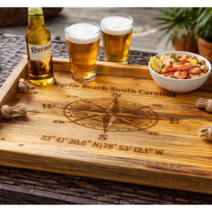 May include: A wooden serving tray with a compass design and the text "Myrtle Beach, South Carolina". The tray holds two glasses of beer, a bottle of Corona beer, and a bowl of shrimp. Decorative rope handles are on the sides.